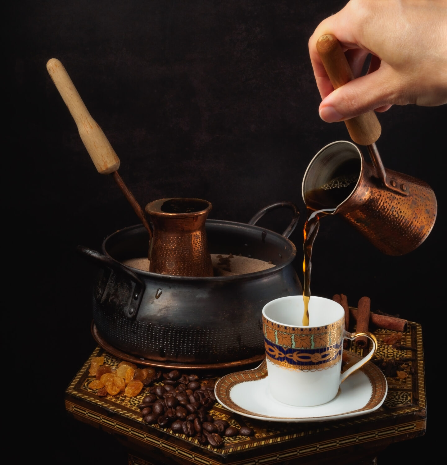 Still life of hot oriental coffee on the sand. Two cezves filled with coffee. One cup of coffee. Female hand pours coffee from turkish into a cup